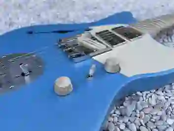 Nahaufnahme einer blauen E-Gitarre mit cremefarbenem Schlagbrett, mehreren Tonabnehmern, Metallbrücke und Holzreglern auf Kies