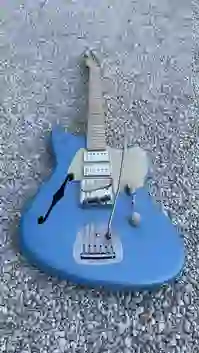 Eine handgefertigte blaue E-Gitarre mit cremefarbenem Schlagbrett und Metallhardware liegt auf hellem Kies