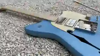 Nahaufnahme des Korpus einer blauen E-Gitarre mit cremefarbenem Schlagbrett, Tonabnehmern, Holzreglern und Metallbrücke auf Kies